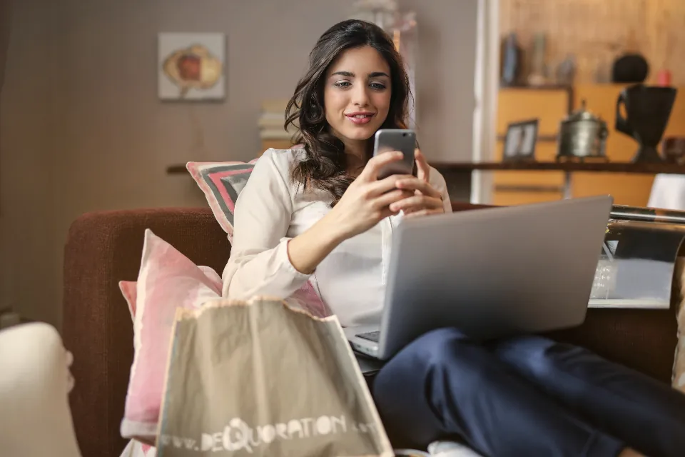 A woman shopping on her phone and laptop with a shopping bag beside her