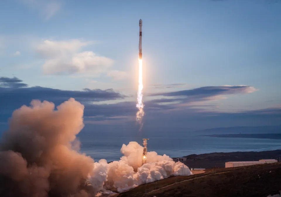 A rocket lifting off from a launch pad into a blue sky with clouds of smoke below