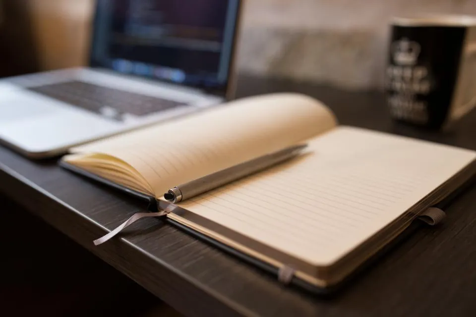 An open notebook in front of a laptop and a coffee mug on a dark desk