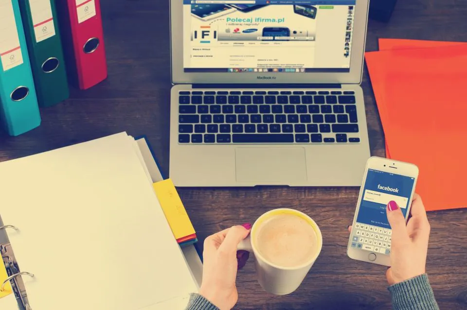 Checking Facebook on a phone beside a laptop and a cup of coffee
